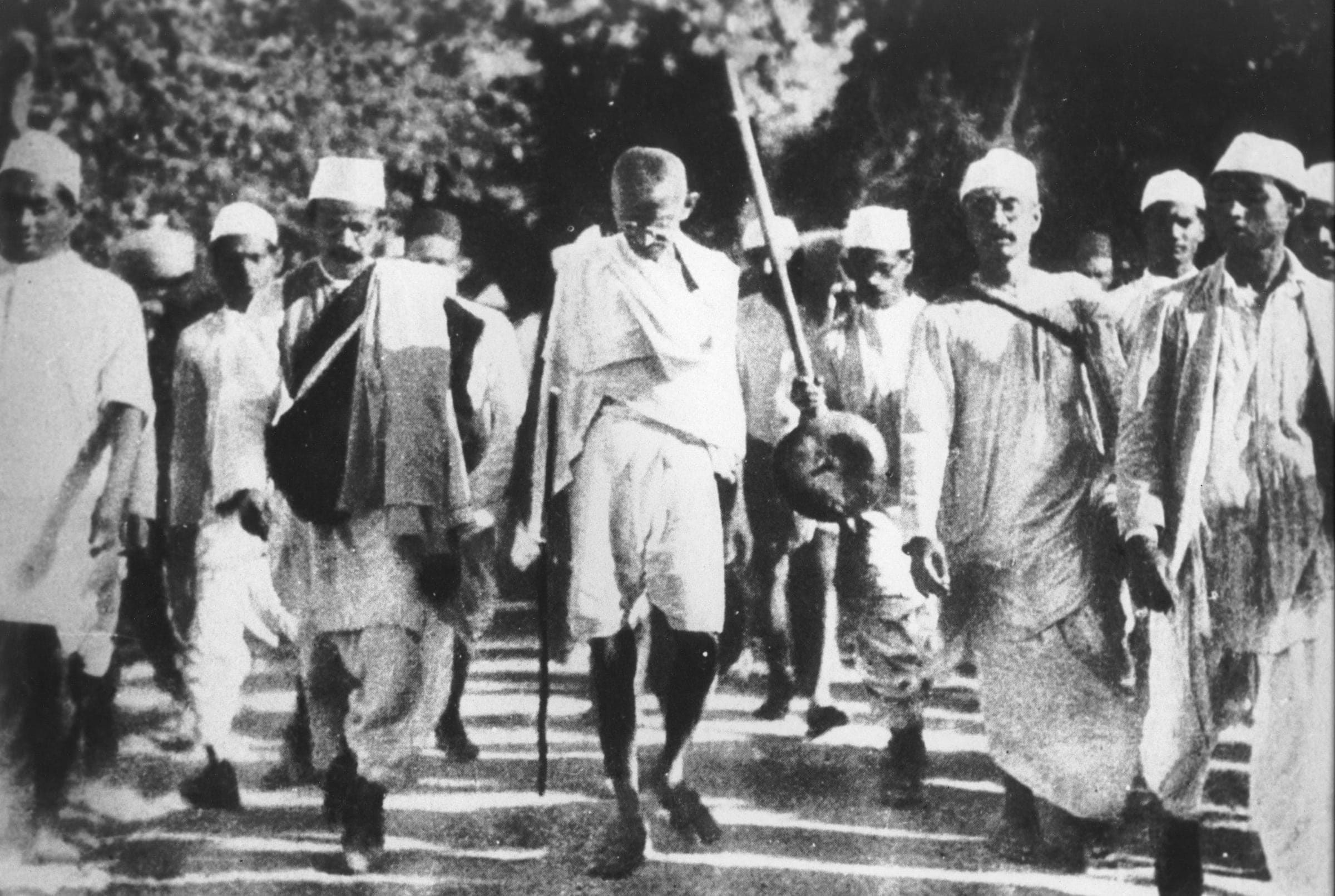 Gandhi, bareheaded, a short walking stick held down at his side, walks at the head of a column of men in white khadi and caps along a tree-shaded road; behind him a man carries a tambura upright.