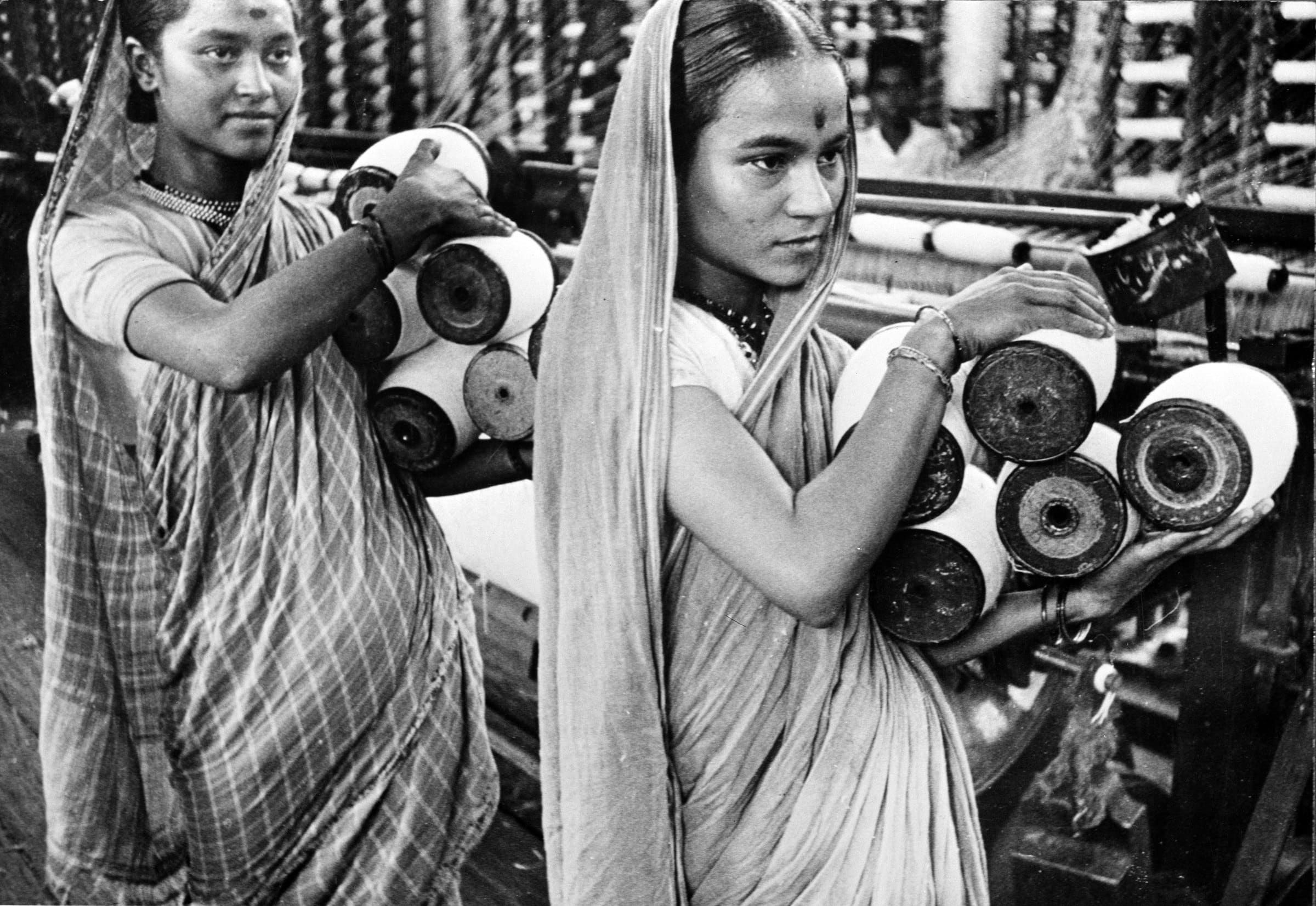 Two young women in checked saris carry stacks of cotton bobbins along a row of spinning machines in a Bombay textile mill; a man stands among the frames behind them.