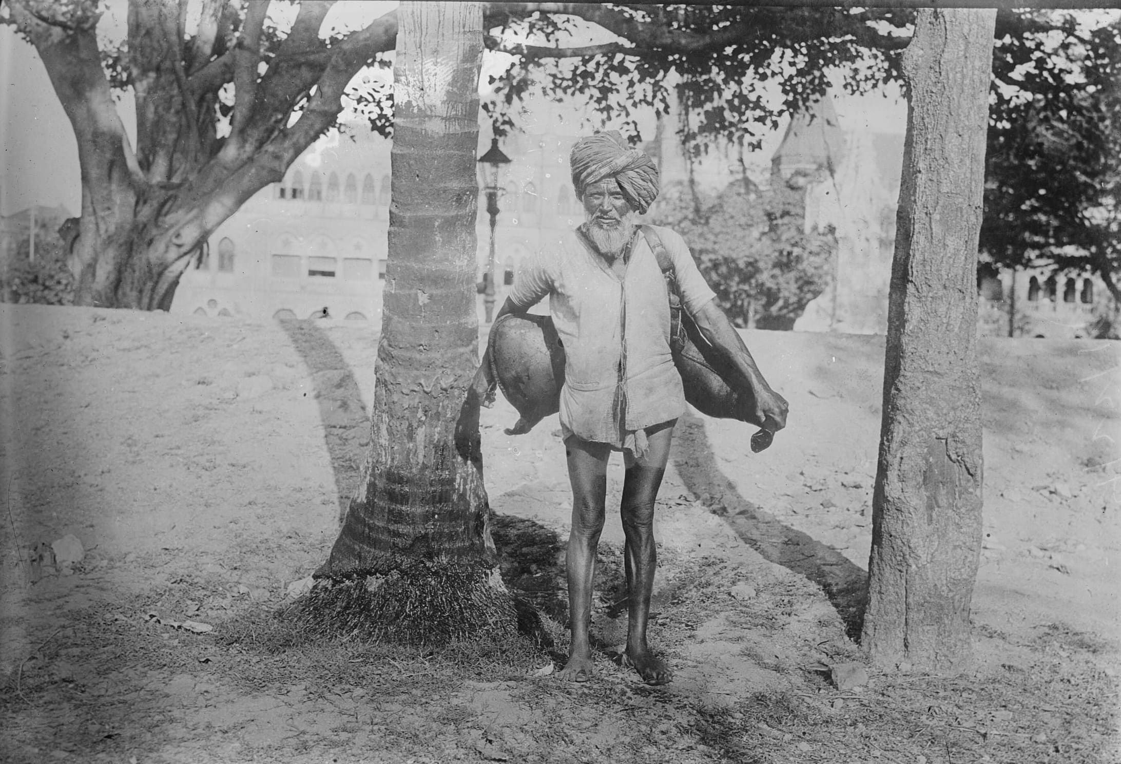 An old man in a turban and a loose cotton shirt, barefoot, stands between two trees on a dusty maidan, a leather water bag slung at his side; behind him, through the trees, are the arches of a large stone building.