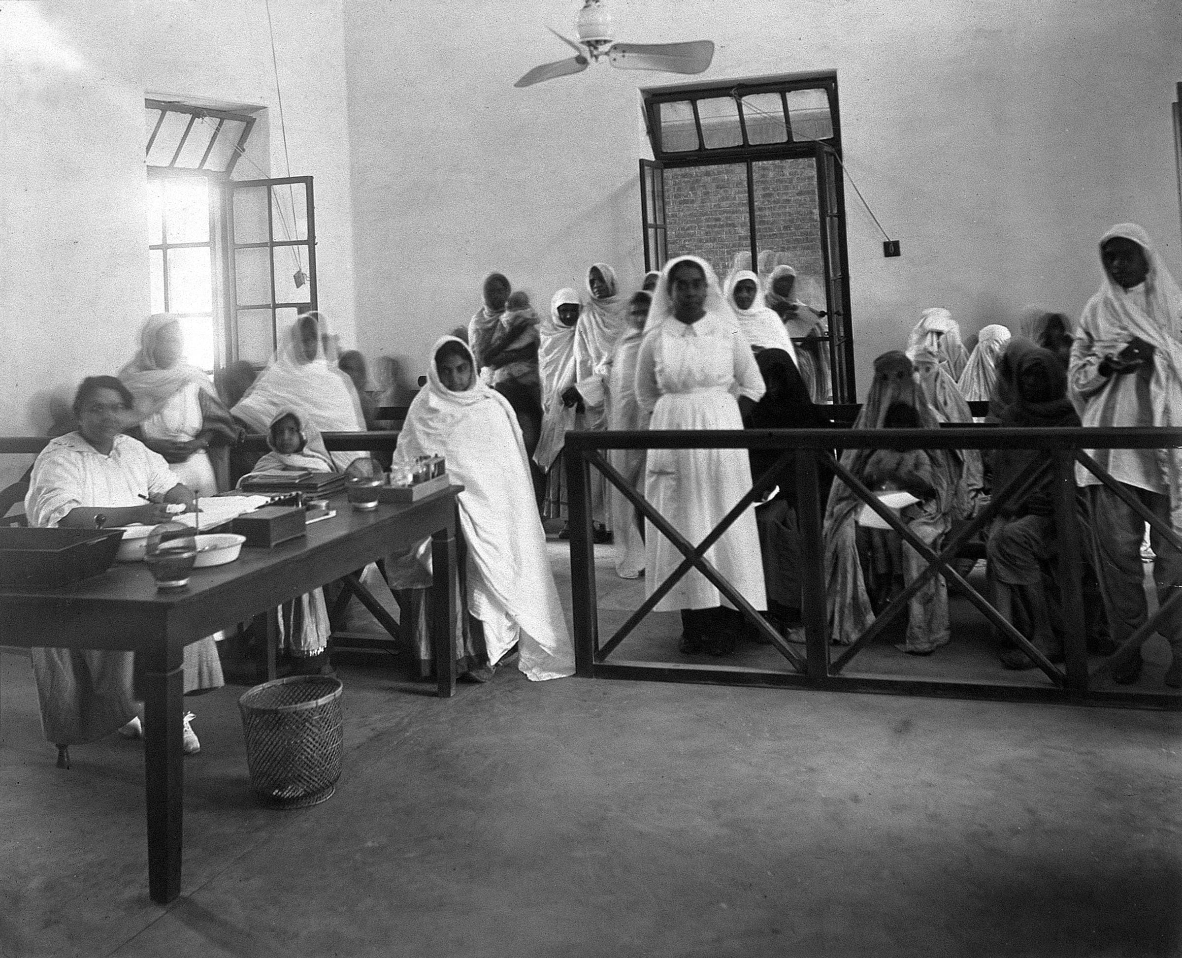 A hospital out-patients' room with whitewashed walls and a ceiling fan. On the left a woman in white sits writing at a table set with bowls and bottles, and a girl in a white shawl waits beside it. Behind a wooden rail, a crowd of women in white wraps, some holding children, stand and sit in line, and a nurse in a white uniform and headscarf stands among them.